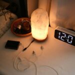 A bedside table just after midnight, with a glowing lamp, a clock showing 12:01 and a device with earphones