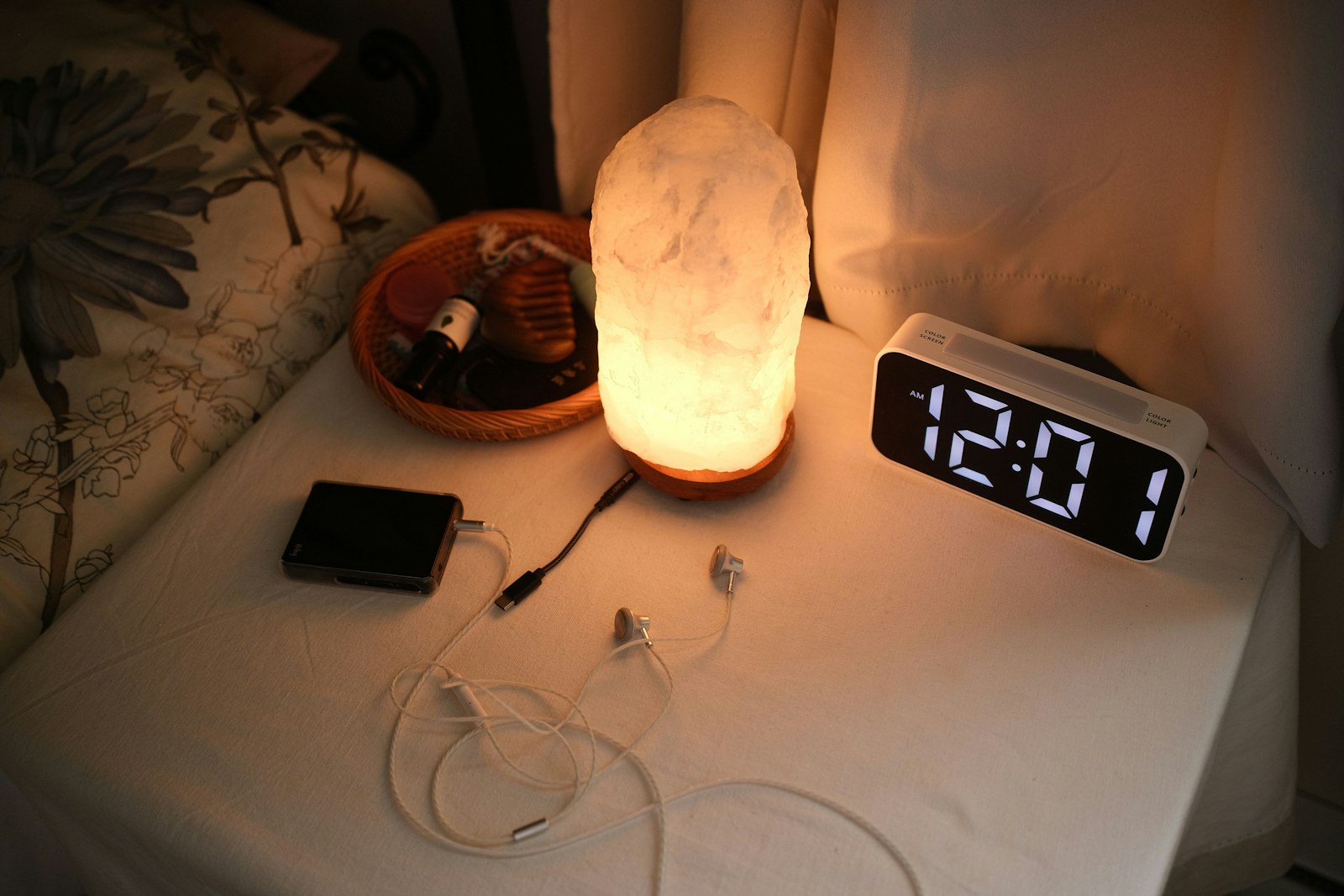 A bedside table just after midnight, with a glowing lamp, a clock showing 12:01 and a device with earphones