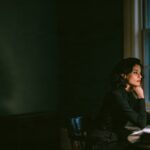 A woman in her late forties sits at a desk by a window at dusk, chin on her hand, lit by a single desk lamp, the rest of the office in shadow
